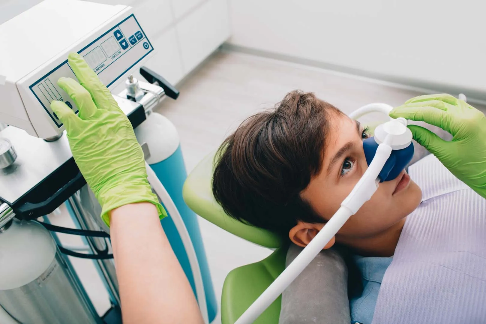 Child lying back with oxygen mask while a gloved hand adjusts the oxygen machine controls.