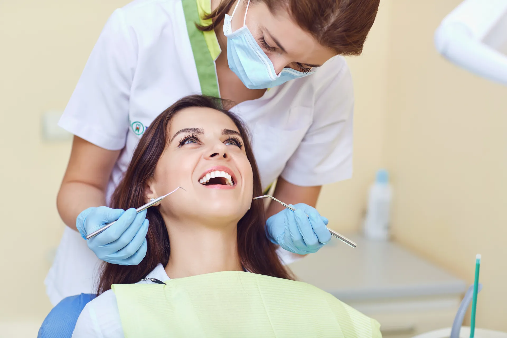 Dentist wearing mask and gloves examines smiling patient's teeth with dental tools in clinic.