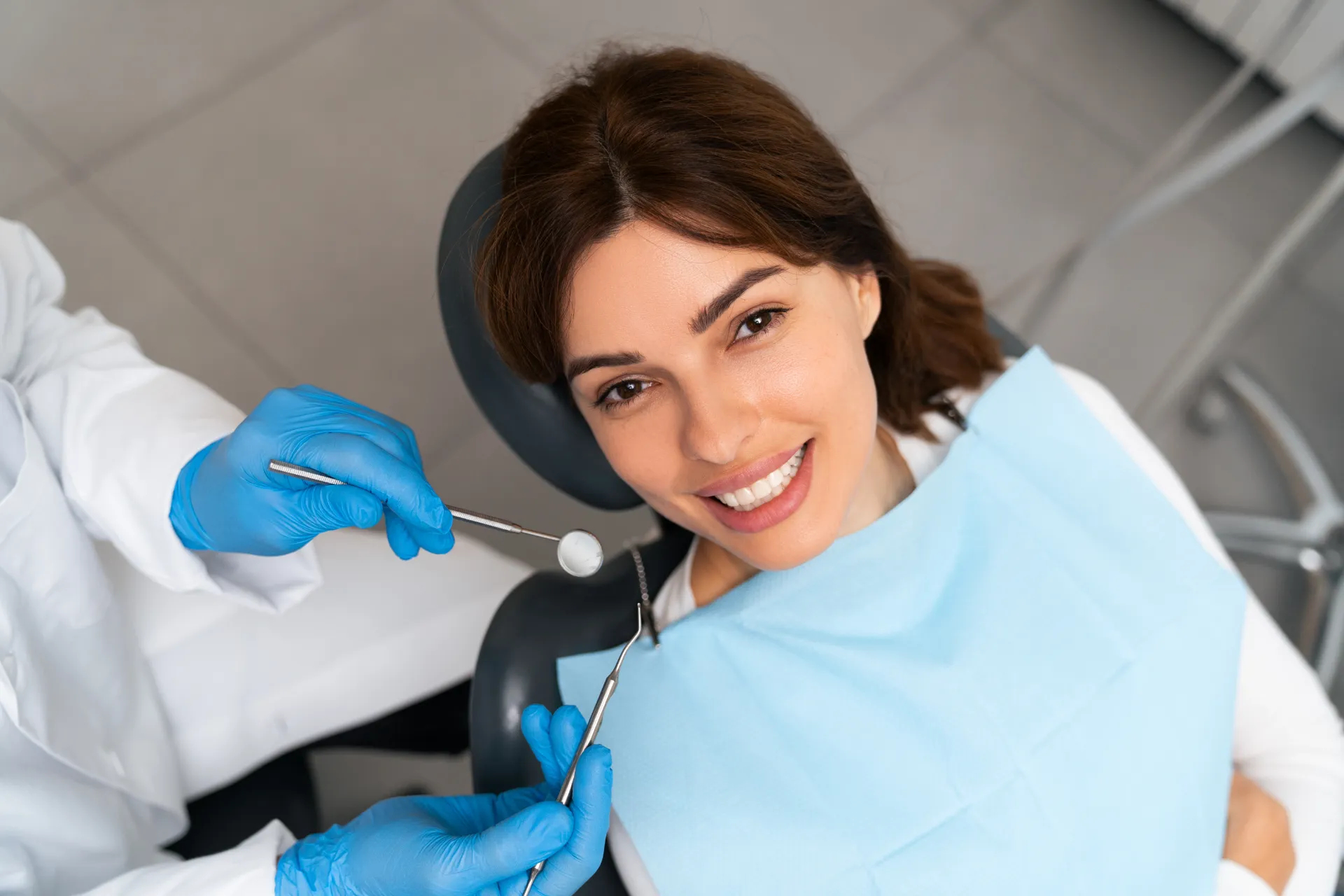 Smiling woman reclined in dental chair with dentist's gloved hands holding dental tools near her mouth.