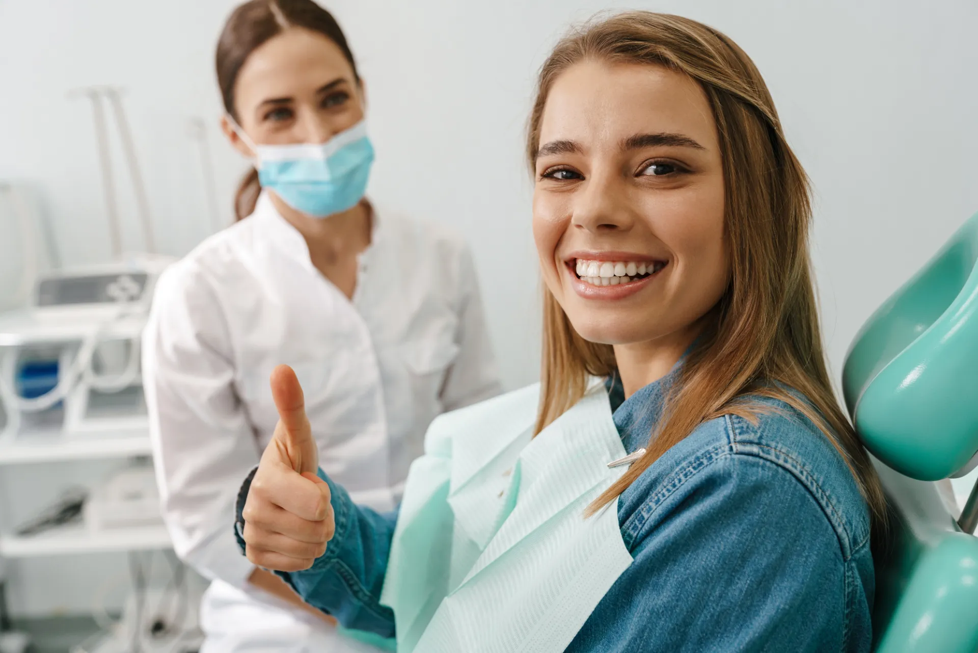 Smiling woman showing thumbs up at dental office with masked dentist in background.