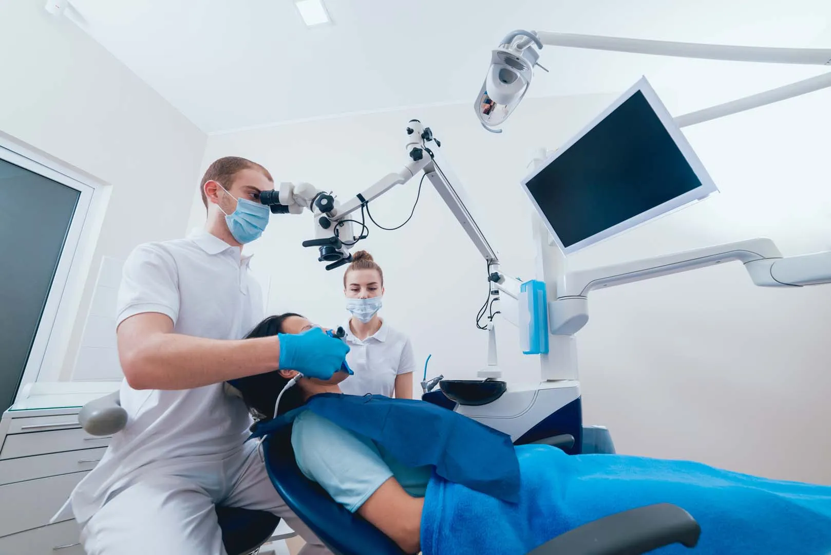 Dentist and assistant wearing masks examining patient using dental microscope in dental clinic.