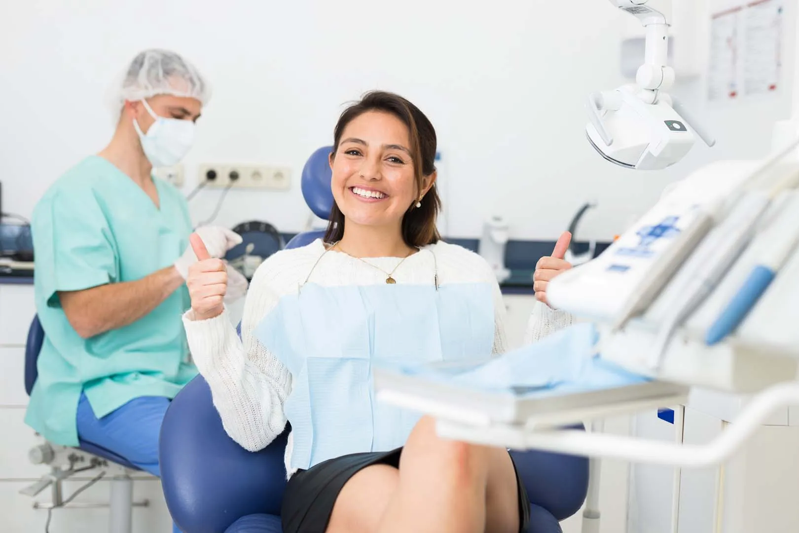 Smiling woman in dental chair giving thumbs up as dentist prepares tools in background.