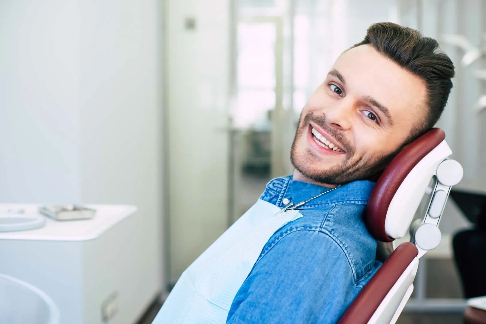 Smiling man in denim shirt sitting in a dental chair with a bib in a dental office.