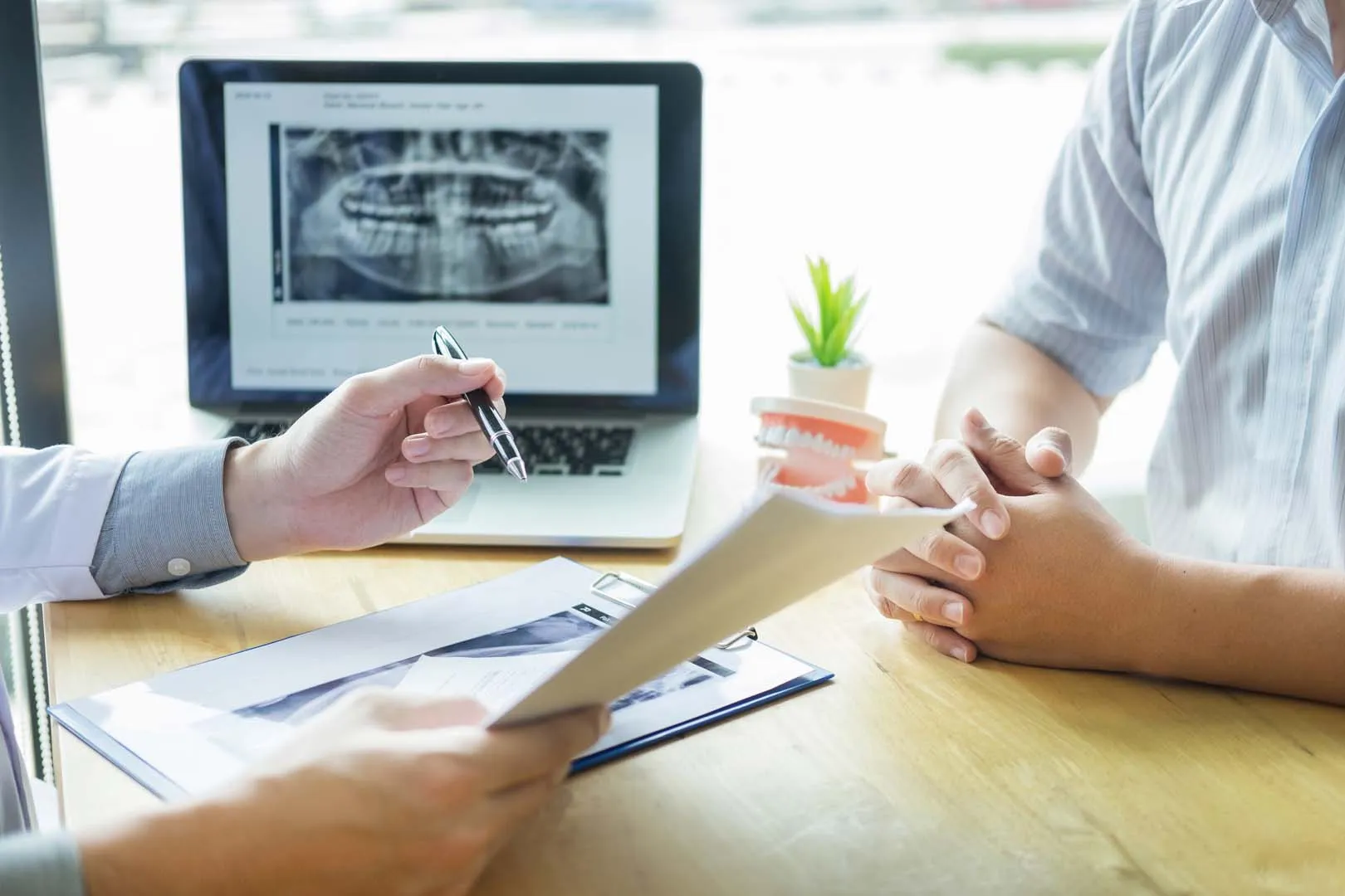 Dentist reviewing dental X-rays on laptop and documents with patient across the table.