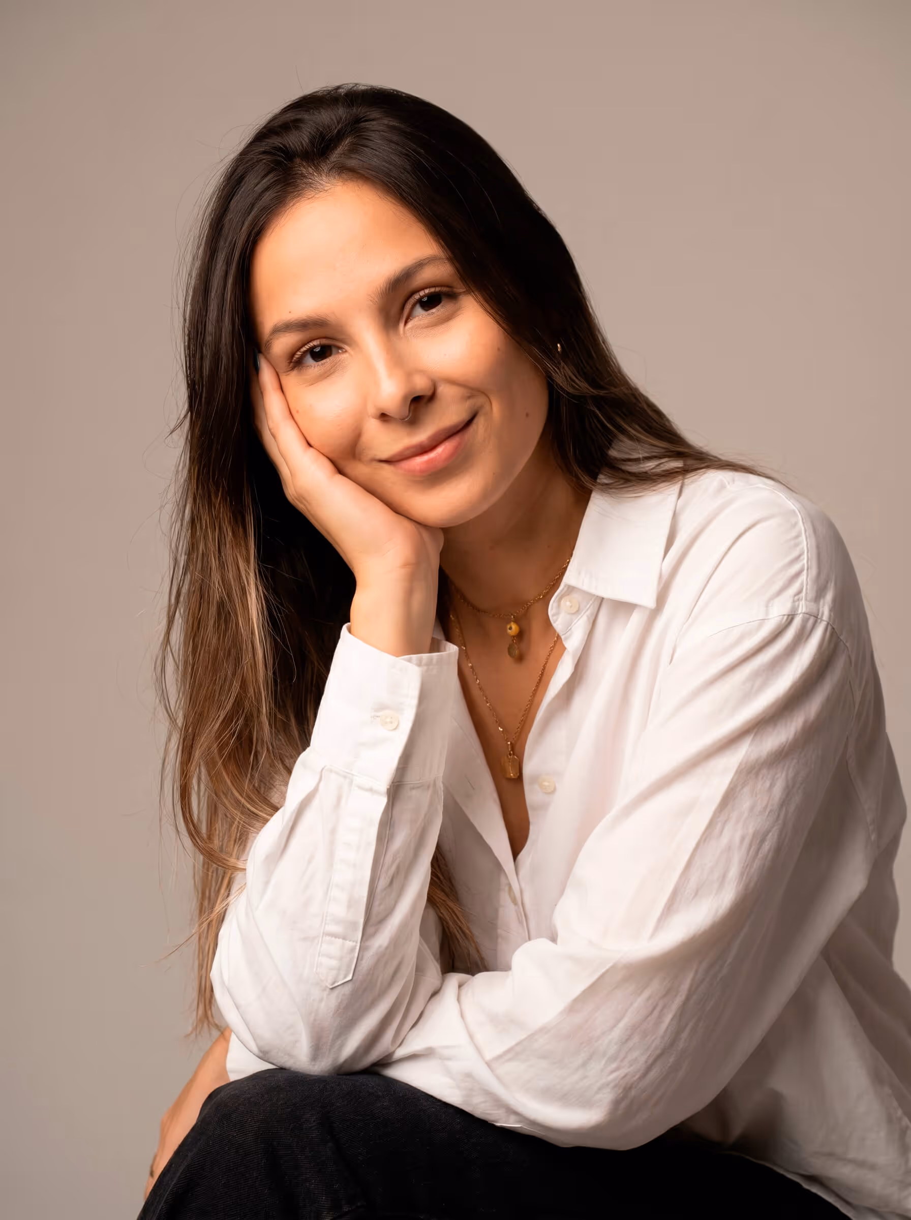 Mujer sonriendo con blusa blanca, apoyando la cabeza en la mano, sobre fondo beige.