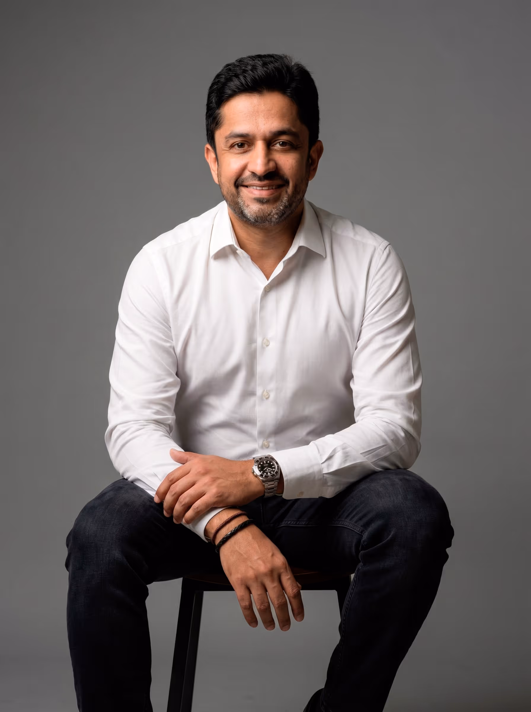 Hombre sonriente con camisa blanca y reloj sentado sobre un taburete con fondo gris.