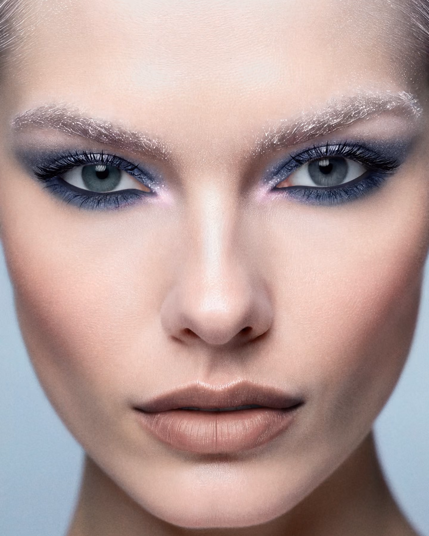 Close-up of a woman's face with frost-coated eyebrows, smoky purple eyeshadow, long dark lashes, gray-green eyes, smooth skin, and nude lipstick.