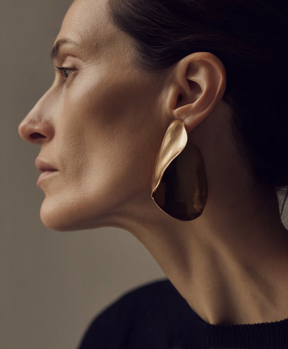 Close-up profile of a woman with dark hair pulled back, wearing a large, abstract-shaped gold earring, against a neutral background.