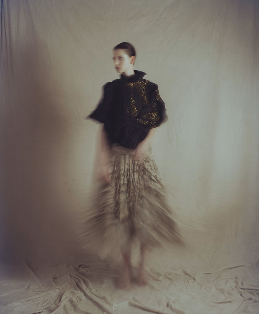 Blurred artistic photo of a person standing barefoot against a beige backdrop, wearing a black top with gold patterns and a beige ruffled skirt, with motion causing a soft, ethereal effect.