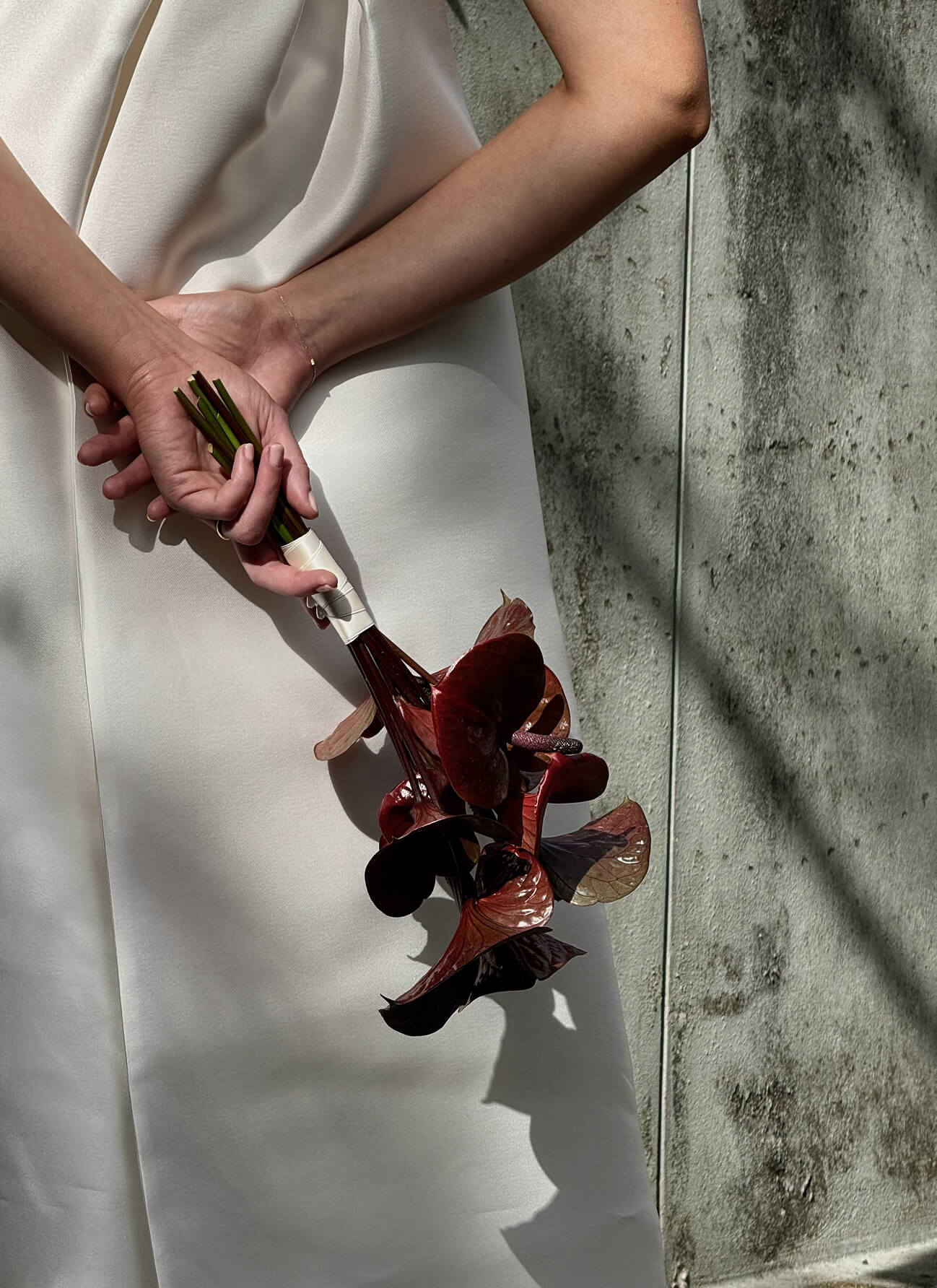 Person in white dress holding a bouquet of dark red calla lilies against a textured wall.
