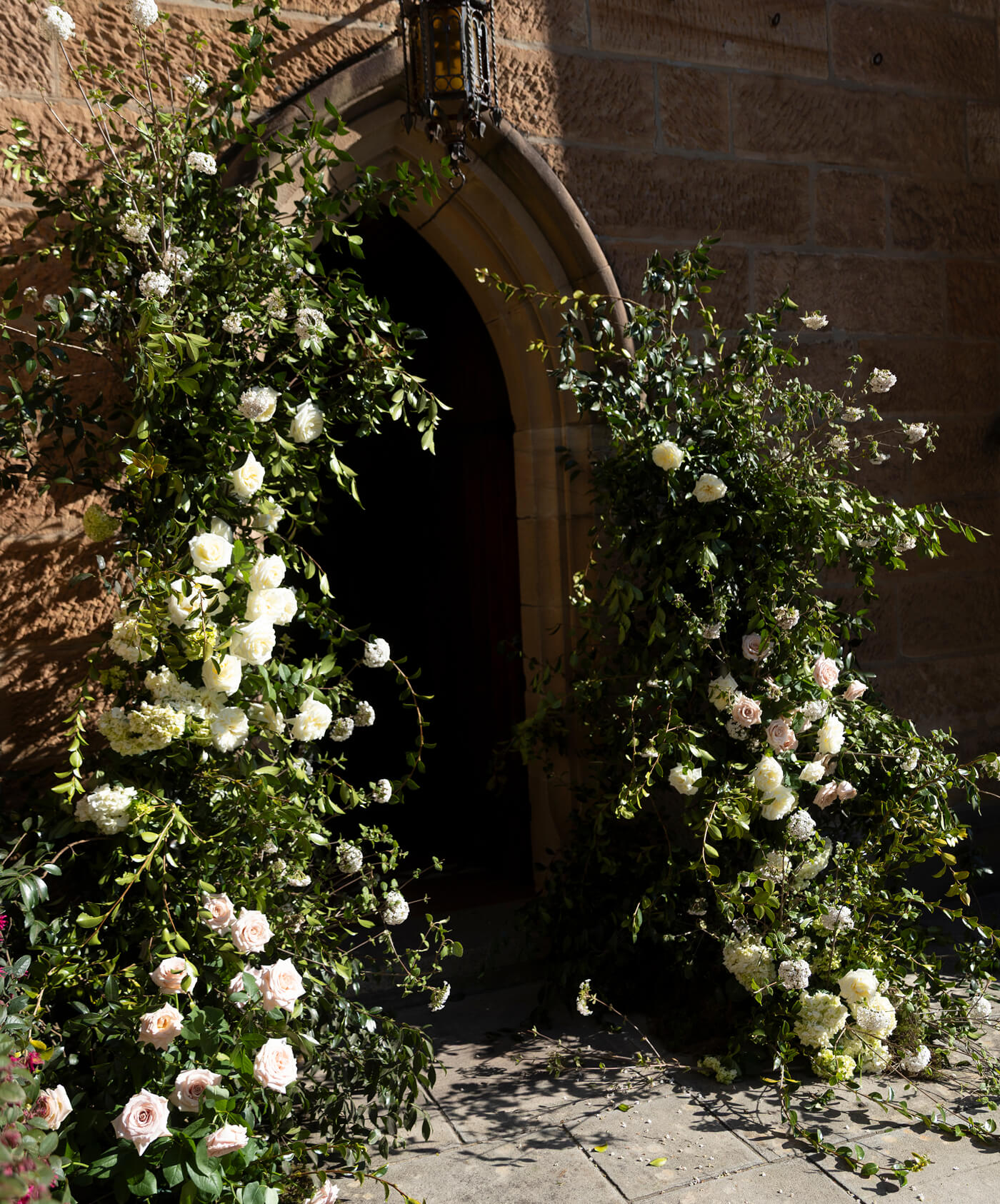 Arched doorway decorated with lush green foliage and white and pale pink roses outside on a stone path.