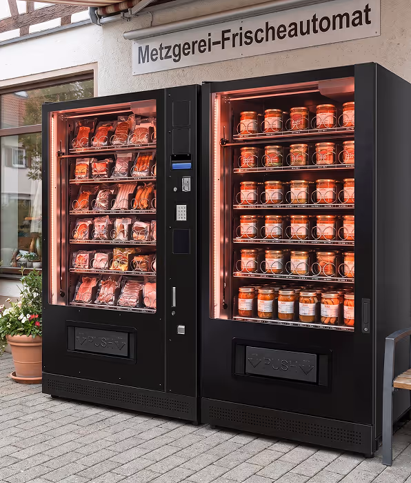 Ein lächelnder junger Mann in einem grauen Anzug und weißem Hemd vor einem städtischen Hintergrund.