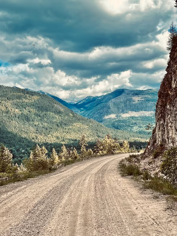 Curved gravel road beside a rocky cliff with forested mountains under a cloudy sky.  Slocan gravel biking
