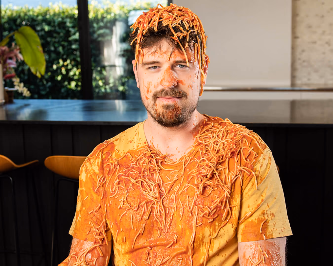 Man with spaghetti and tomato sauce covering his head, face, and orange shirt, sitting indoors.