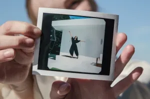 Person holding a Polaroid photo showing a woman jumping with arms raised outdoors against a white wall.