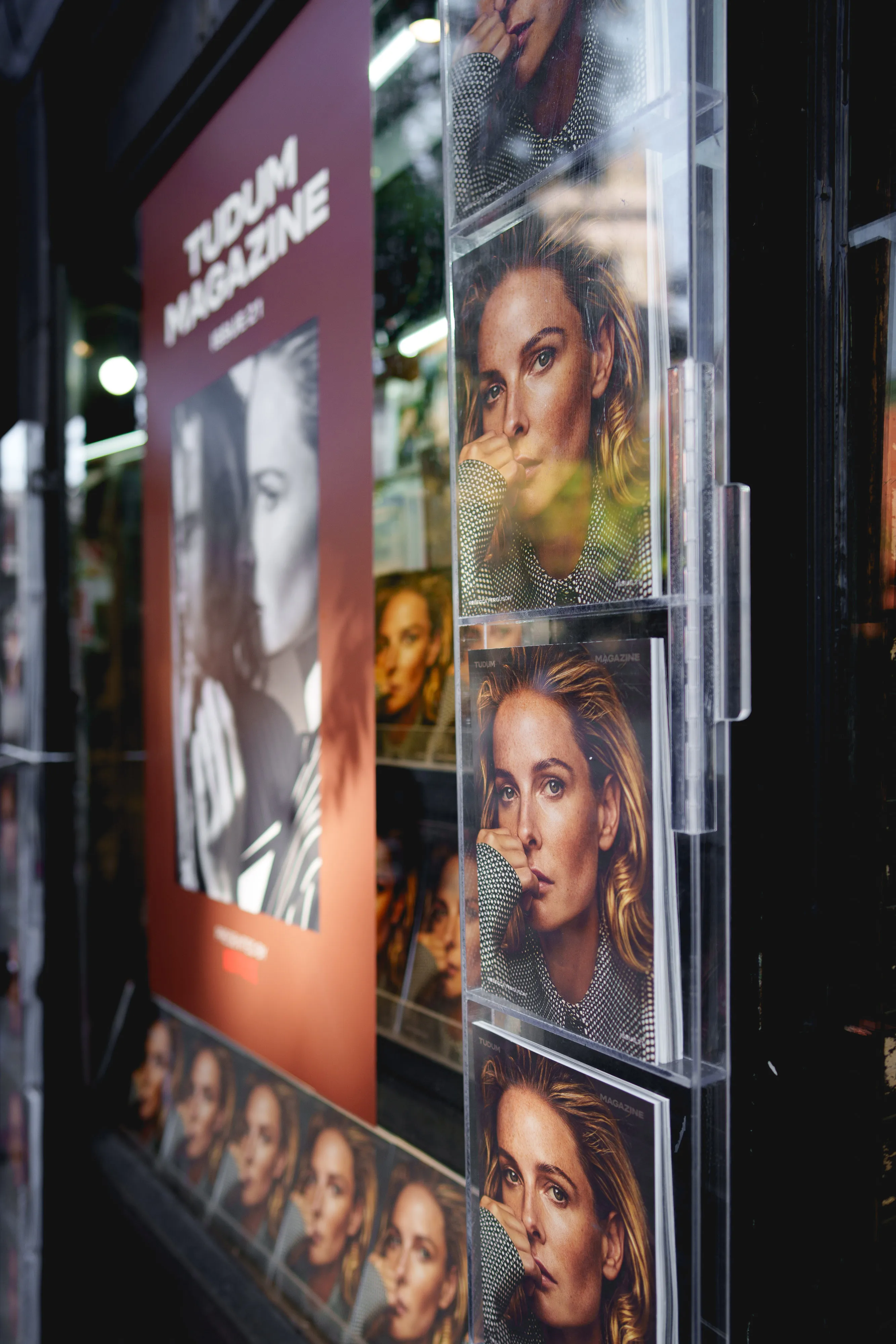 Copies of TUDUM magazine showing a close-up of a woman resting her chin on her hand displayed outside a store.