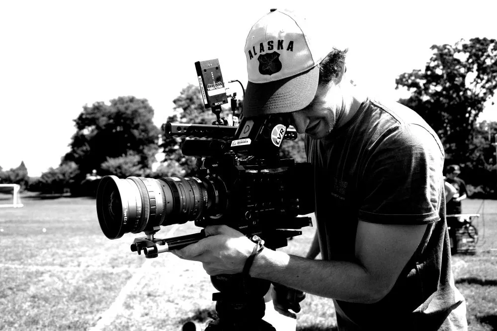 Man wearing an Alaska cap operating a professional video camera outdoors on a sunny day.