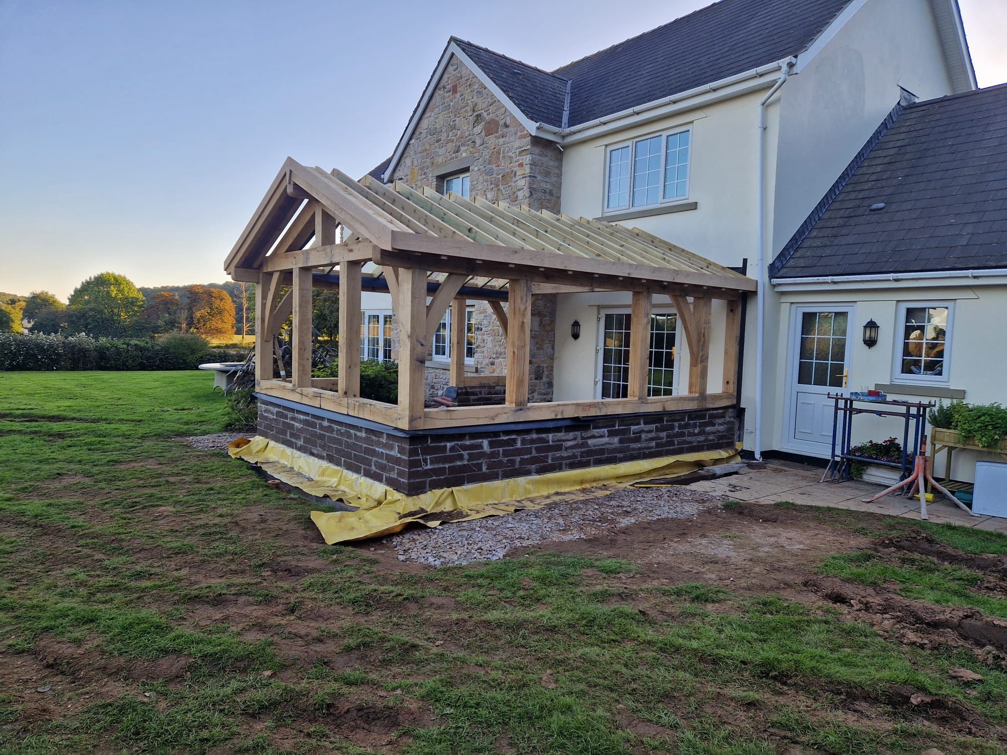 A newly raised oak frame extension standing on its brick base against a house at Caerwent, with the rafters on.