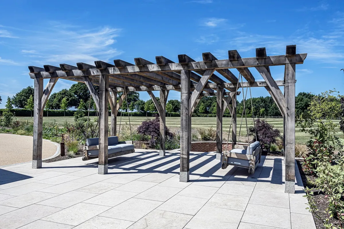 The oak pergola at Ullenhall, Warwickshire, with two hanging oak sofas under it on a paved terrace