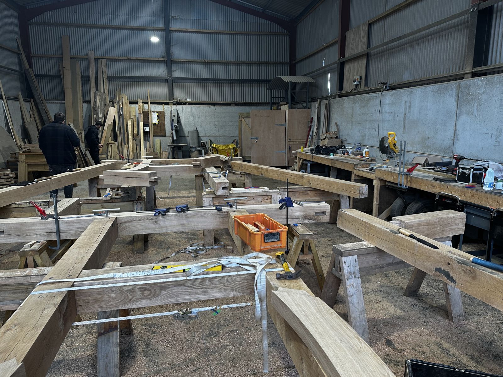 Inside the Foxtail Oak workshop: oak frame timbers on trestles, clamped and strapped, with two of the team at work
