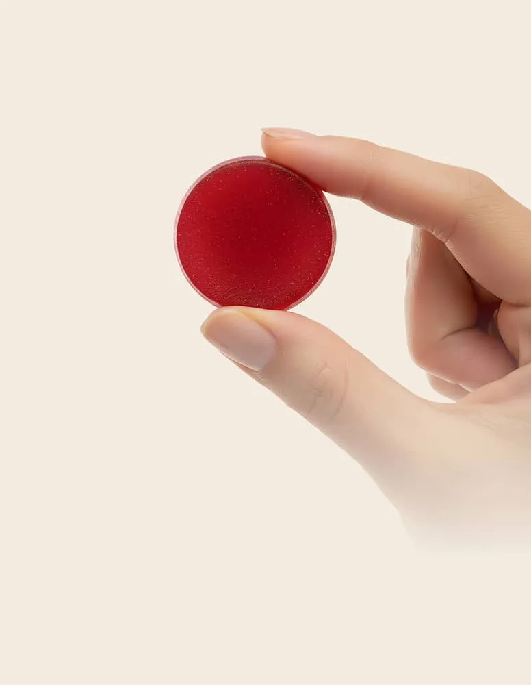 Hand holding a round red solid object between thumb and forefinger against a light background.