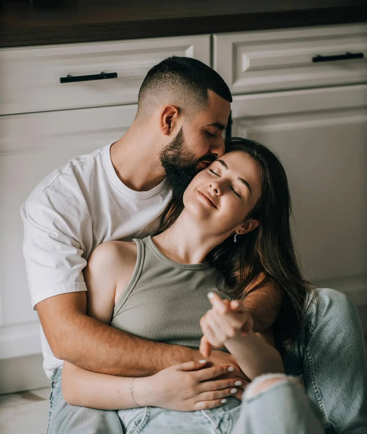 Couple sitting close, man hugging woman and kissing her forehead as she smiles with eyes closed.
