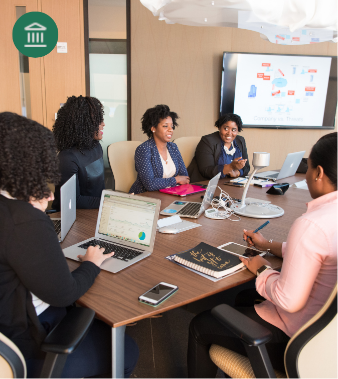 Five women sit around a conference table with laptops and notebooks in a business meeting room. Illustrating Decision Makers of Institutions like HMOs, NGOs, Banks, Employers enjoying Outpatient Health Access programs for customers, staff and communities