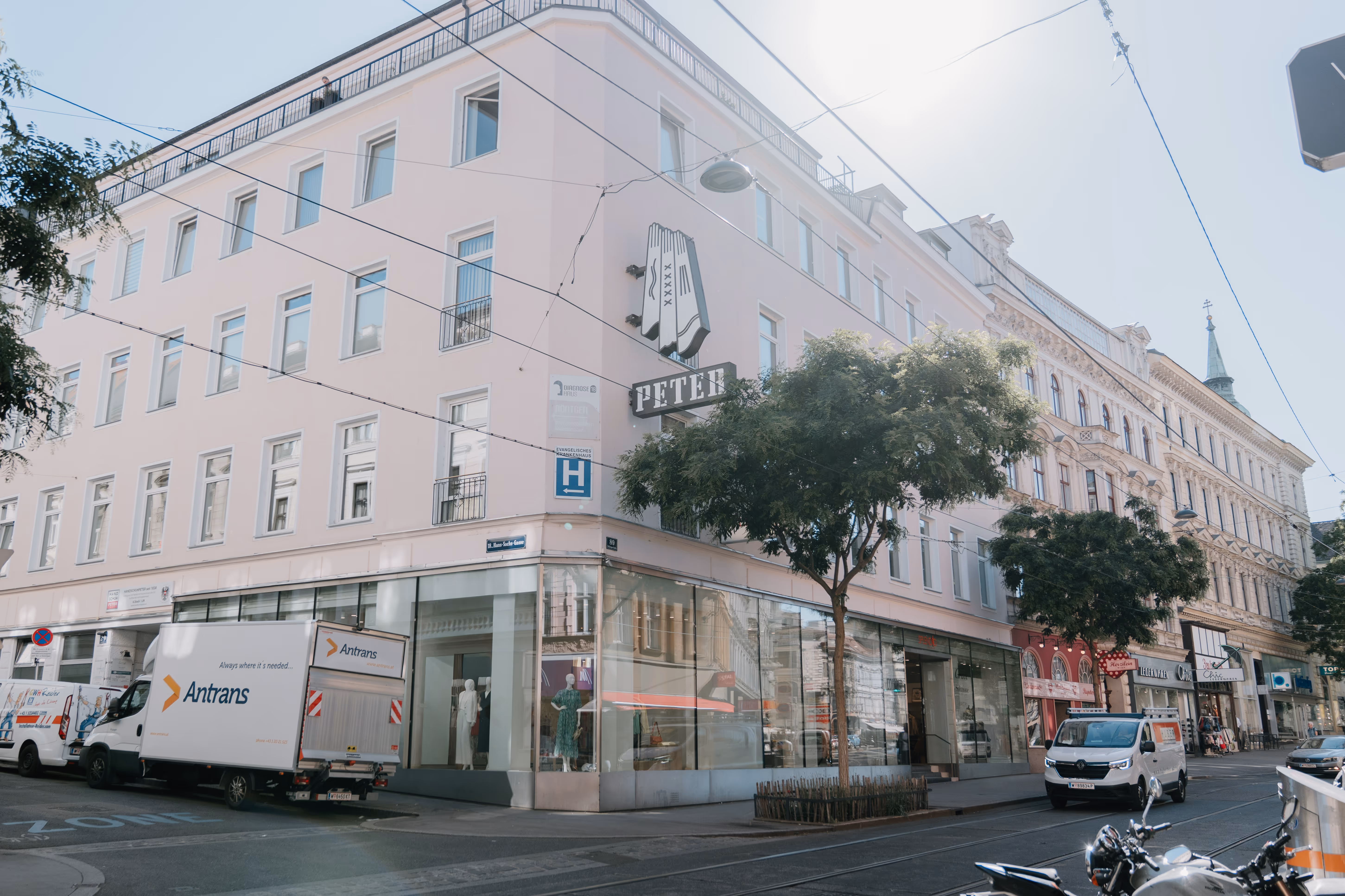Straßenecke mit einem Gebäude, Bäumen, parkenden Lieferwagen und Schaufenstern eines Bekleidungsgeschäfts.