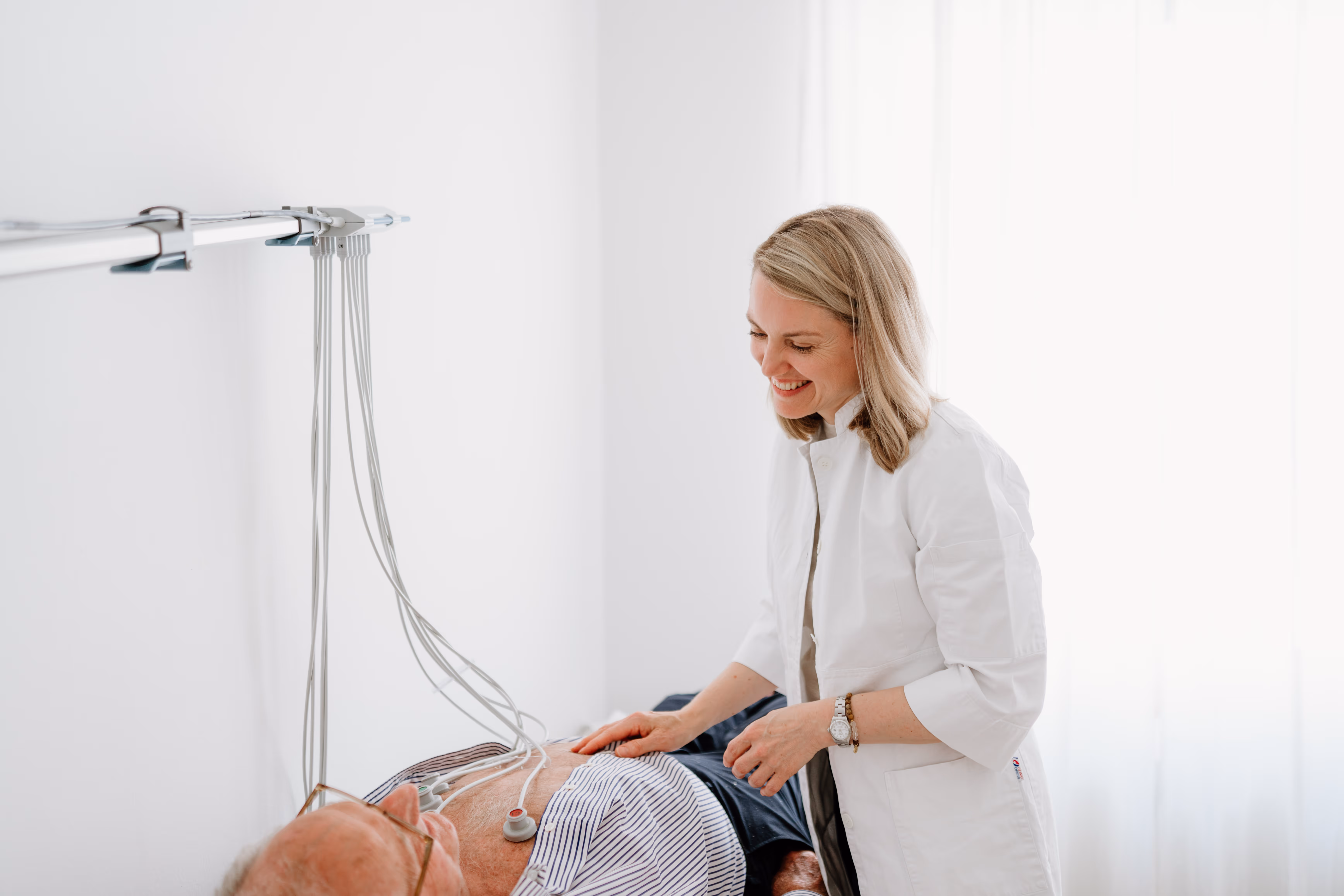 Ärztin führt EKG bei einem älteren, liegenden Mann in heller Praxis durch.