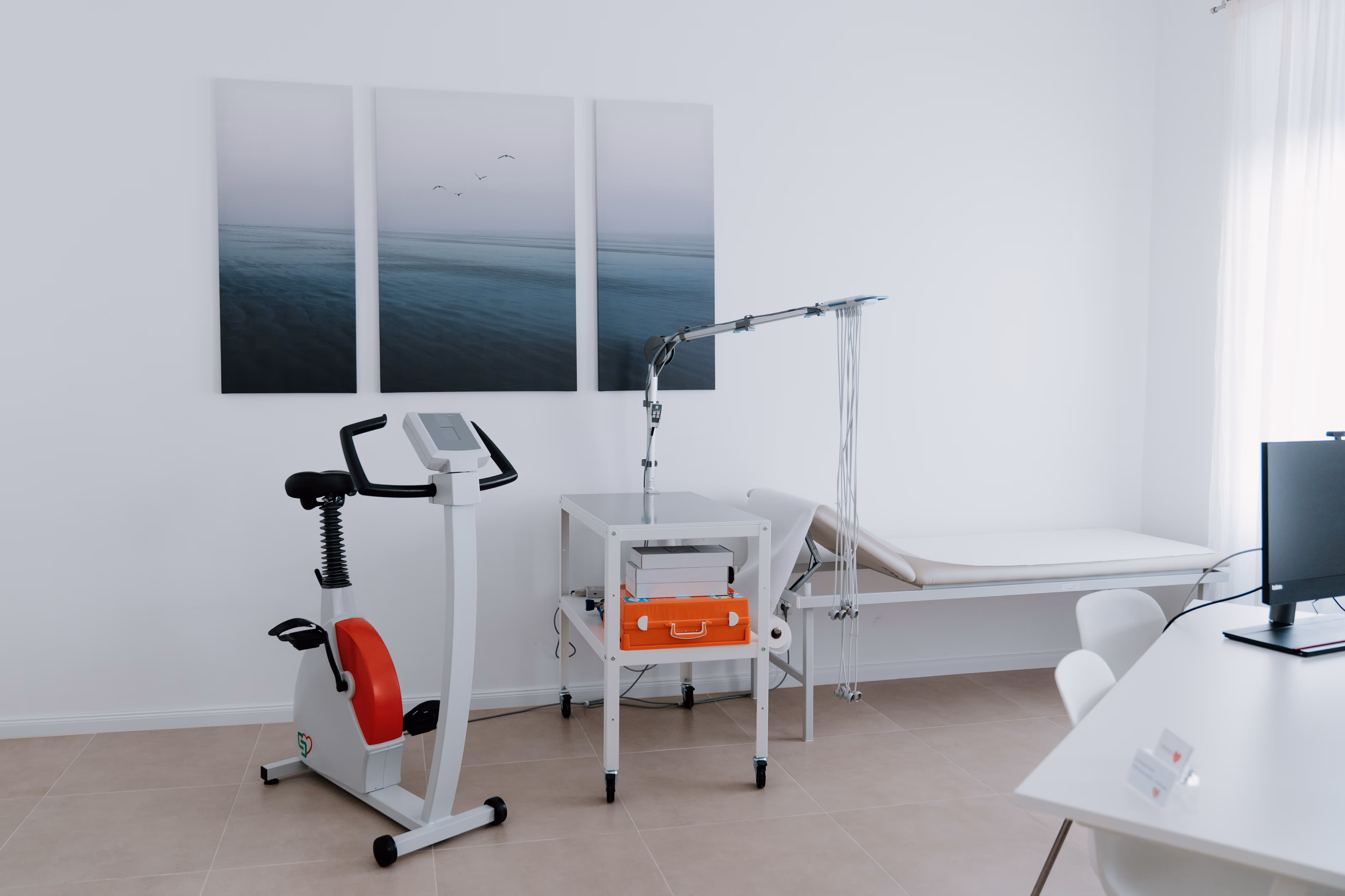 Klinikraum mit Heimtrainer, medizinischer Ausrüstung, Liege und Schreibtisch mit Computer.