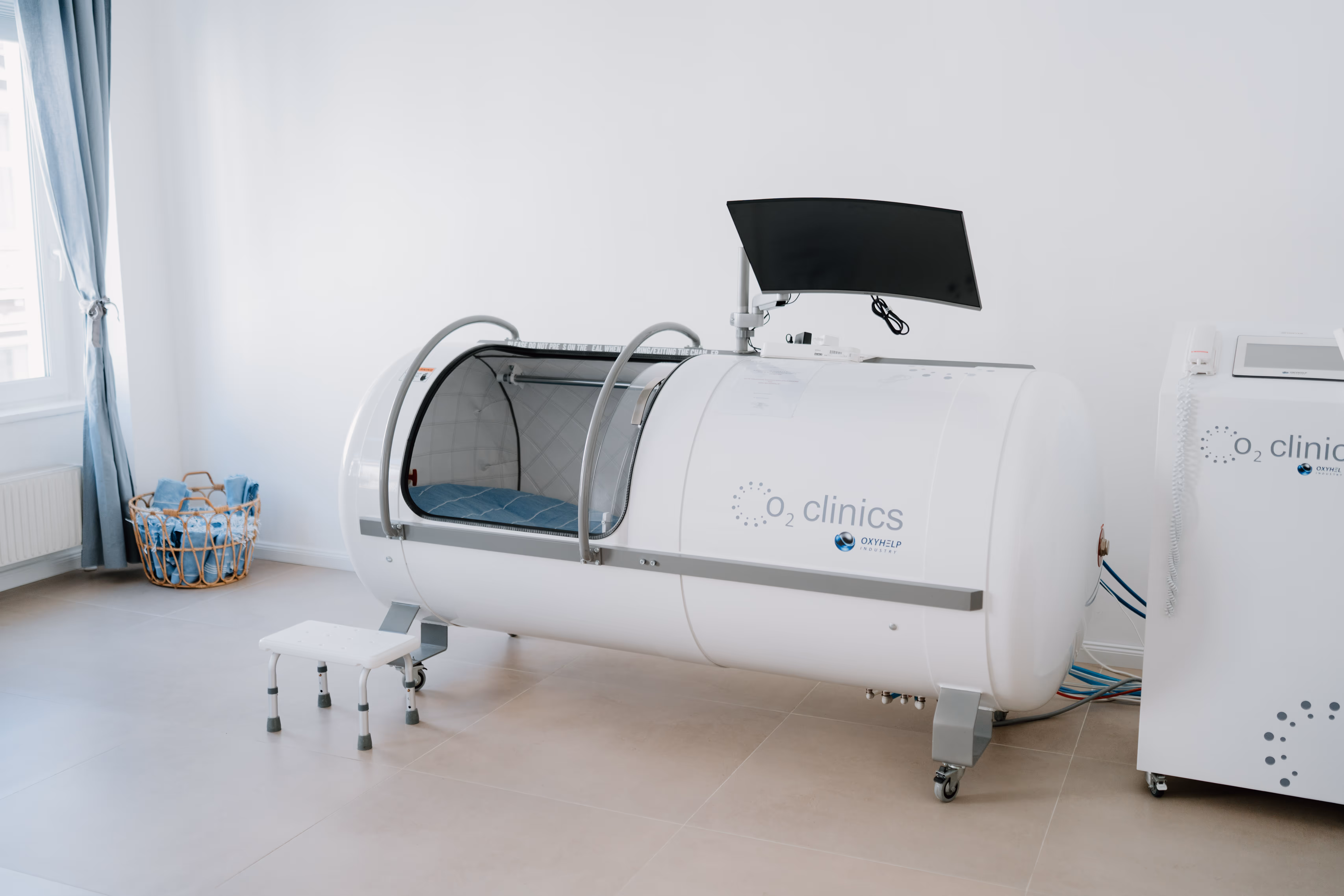 Weiße Hyperbarkammer mit geöffnetem transparentem Deckel, Monitor und kleiner Trittleiter in Klinikraum.