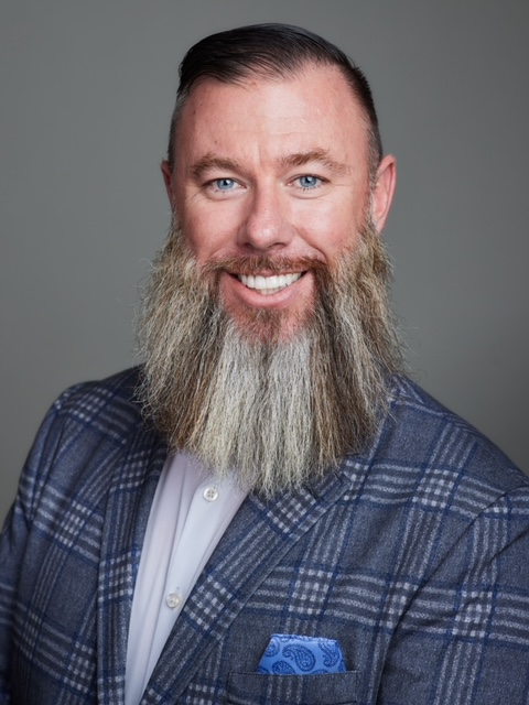Smiling man with long gray beard wearing a blue plaid blazer and white shirt against gray background.