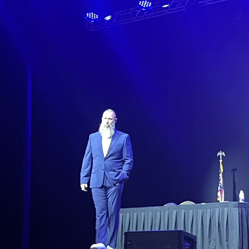 Man with a white beard in a blue suit standing on a stage with a table and American flag behind him.