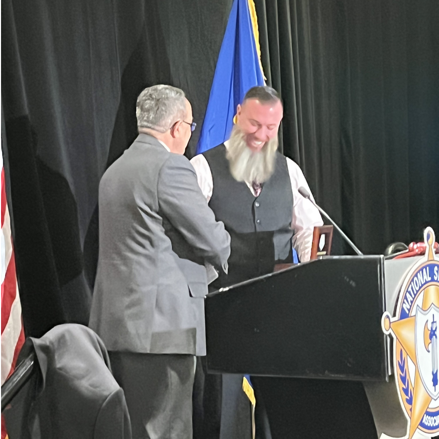 Two men shaking hands at a podium with a National Sheriff's Association emblem in the background.