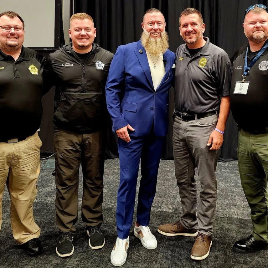 Five men standing and smiling, including four in law enforcement attire and one in a blue suit.