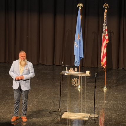 Man with beard in light blue blazer stands near podium with flags on stage with dark curtains.