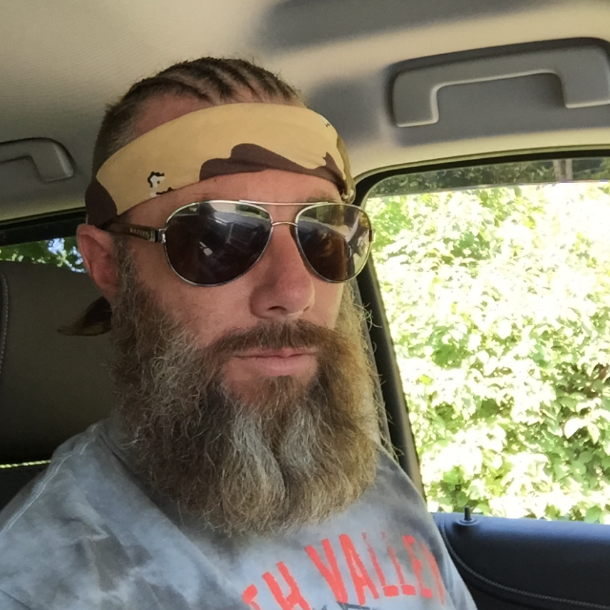 Bearded man wearing sunglasses and a camo headband sitting inside a car with greenery outside.