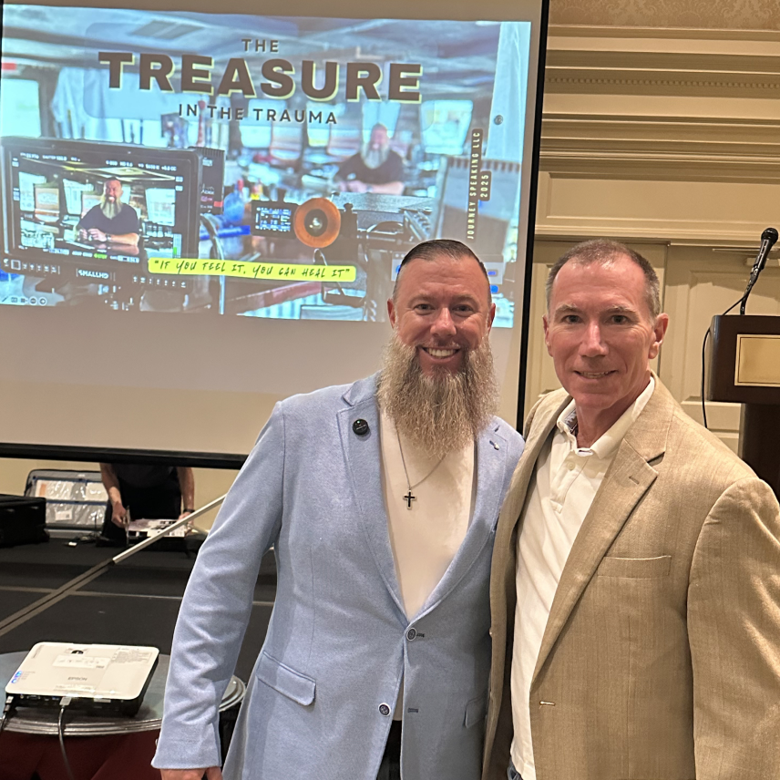 Two men smiling in front of a screen displaying 'The Treasure in the Trauma' presentation.