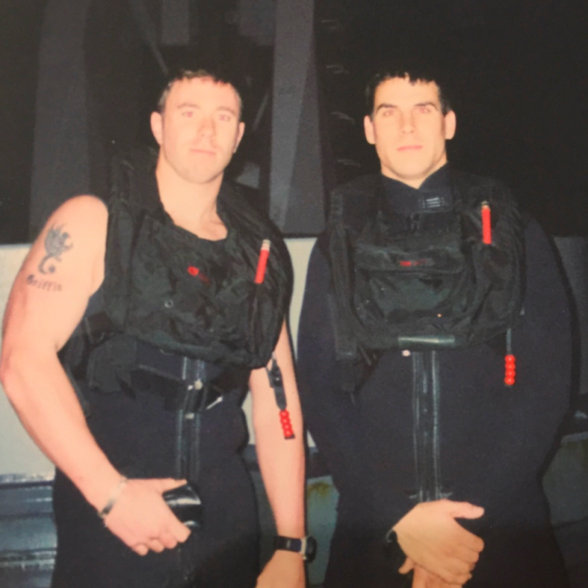 Two men wearing black wetsuits and diving vests standing side by side in dim light.