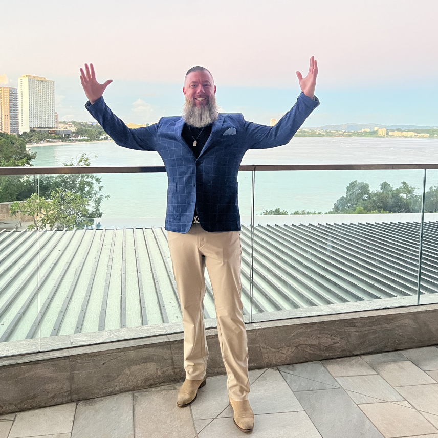 Bearded man in blue jacket and beige pants stands with arms raised on a balcony overlooking water.