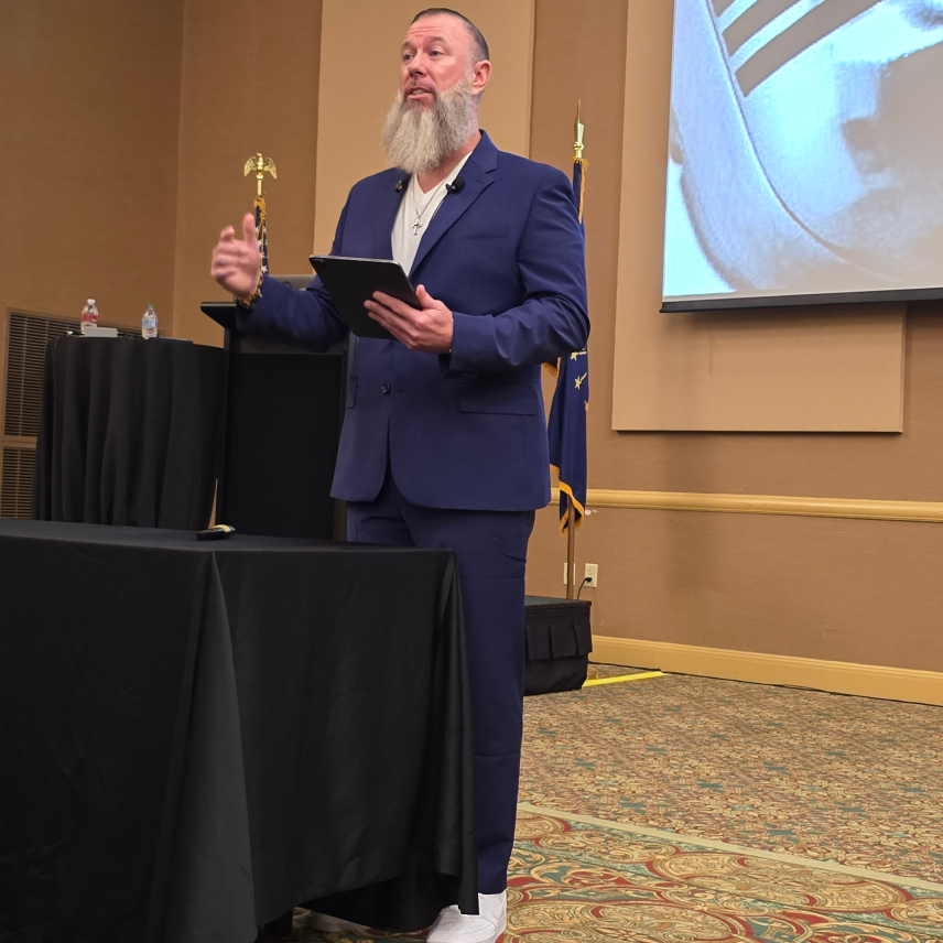 Bearded man in blue suit holding tablet speaking at an event with flags and screen behind him.