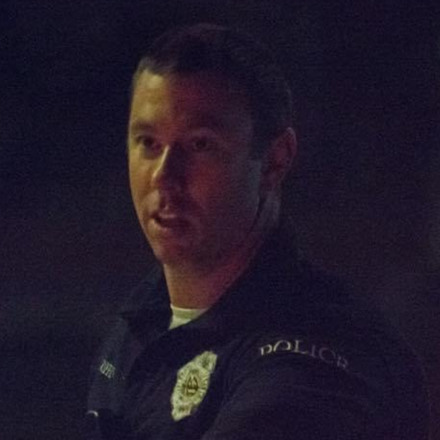Police officer in uniform shown from the chest up against a dark background at night.