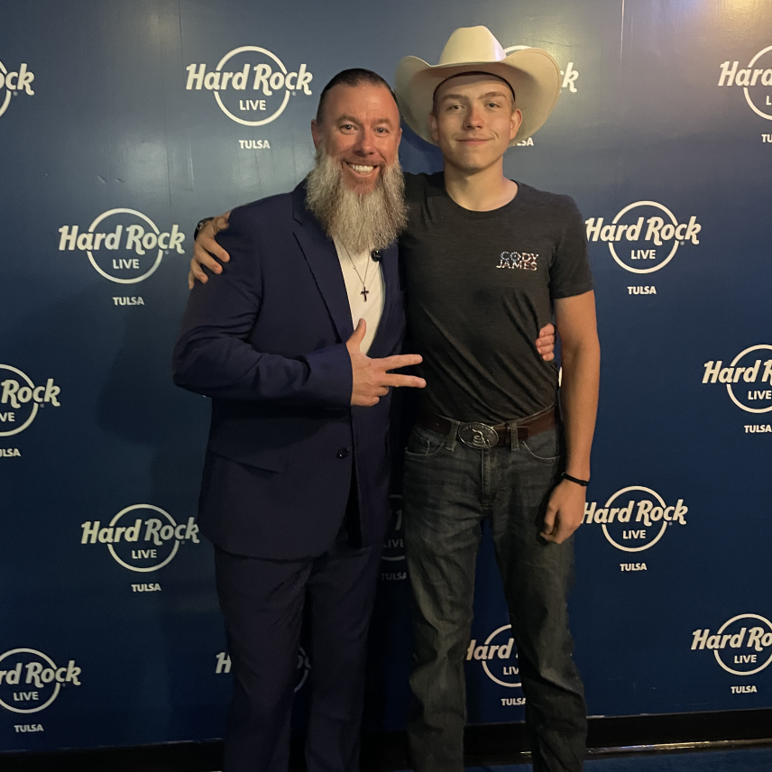 Two men smiling in front of a Hard Rock Live Tulsa backdrop, one wearing a suit, the other a cowboy hat.