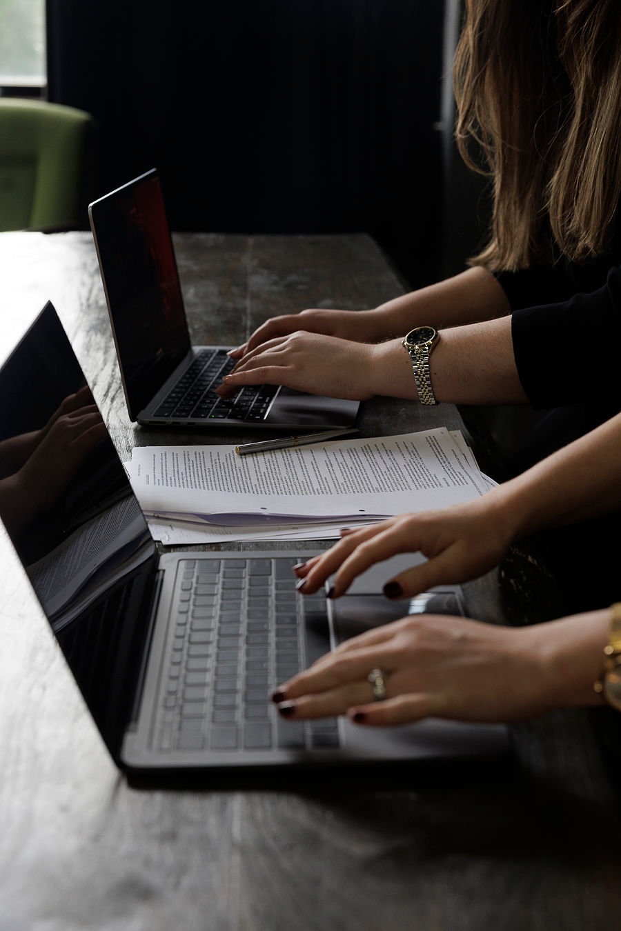 Duas mulheres a trabalhar em laptops com documentos e uma caneta numa mesa de madeira escura.