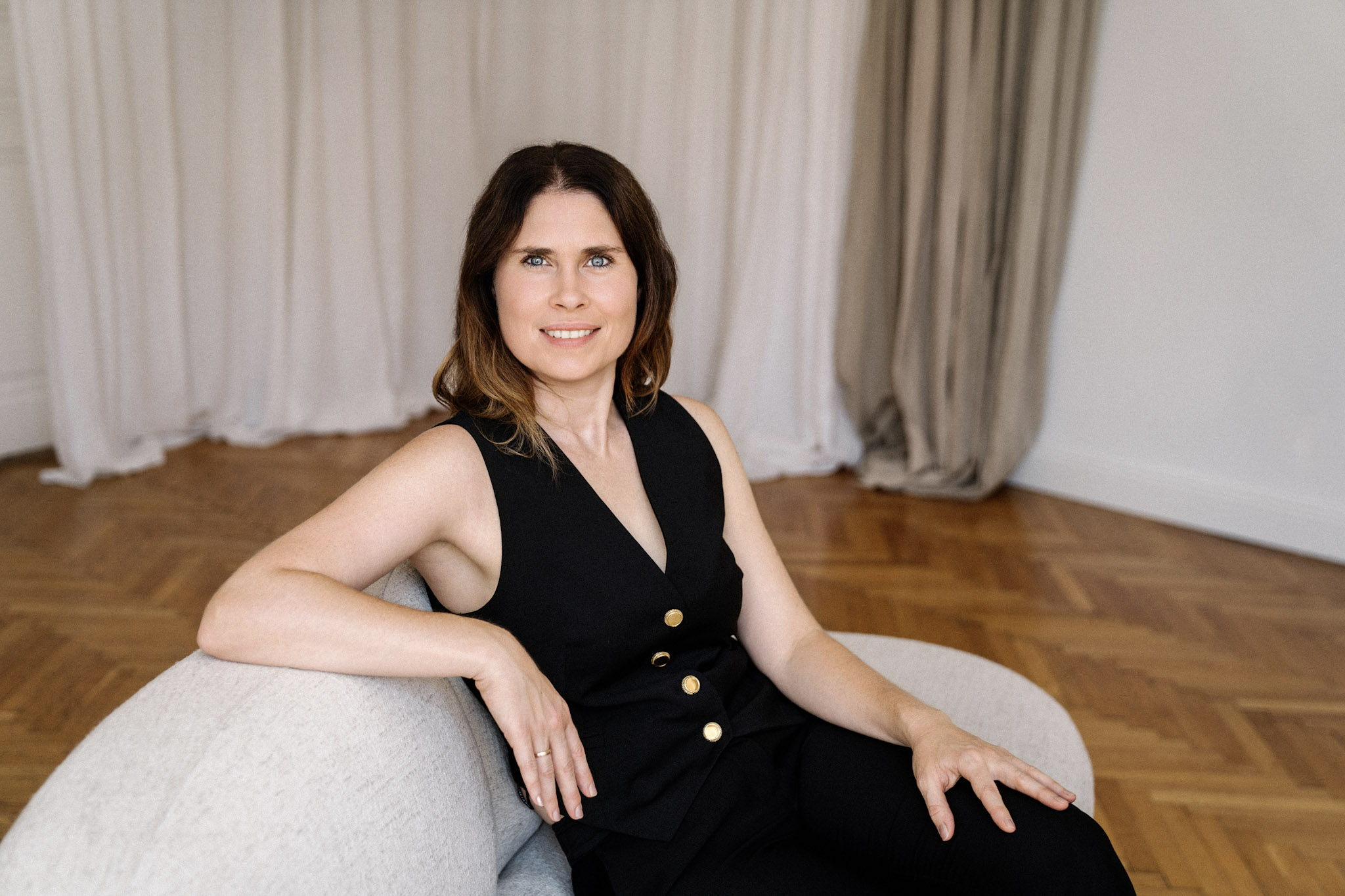 Christina Stix-Pusswald sitzt lächelnd auf grauem Sofa vor cremefarbenen Vorhängen