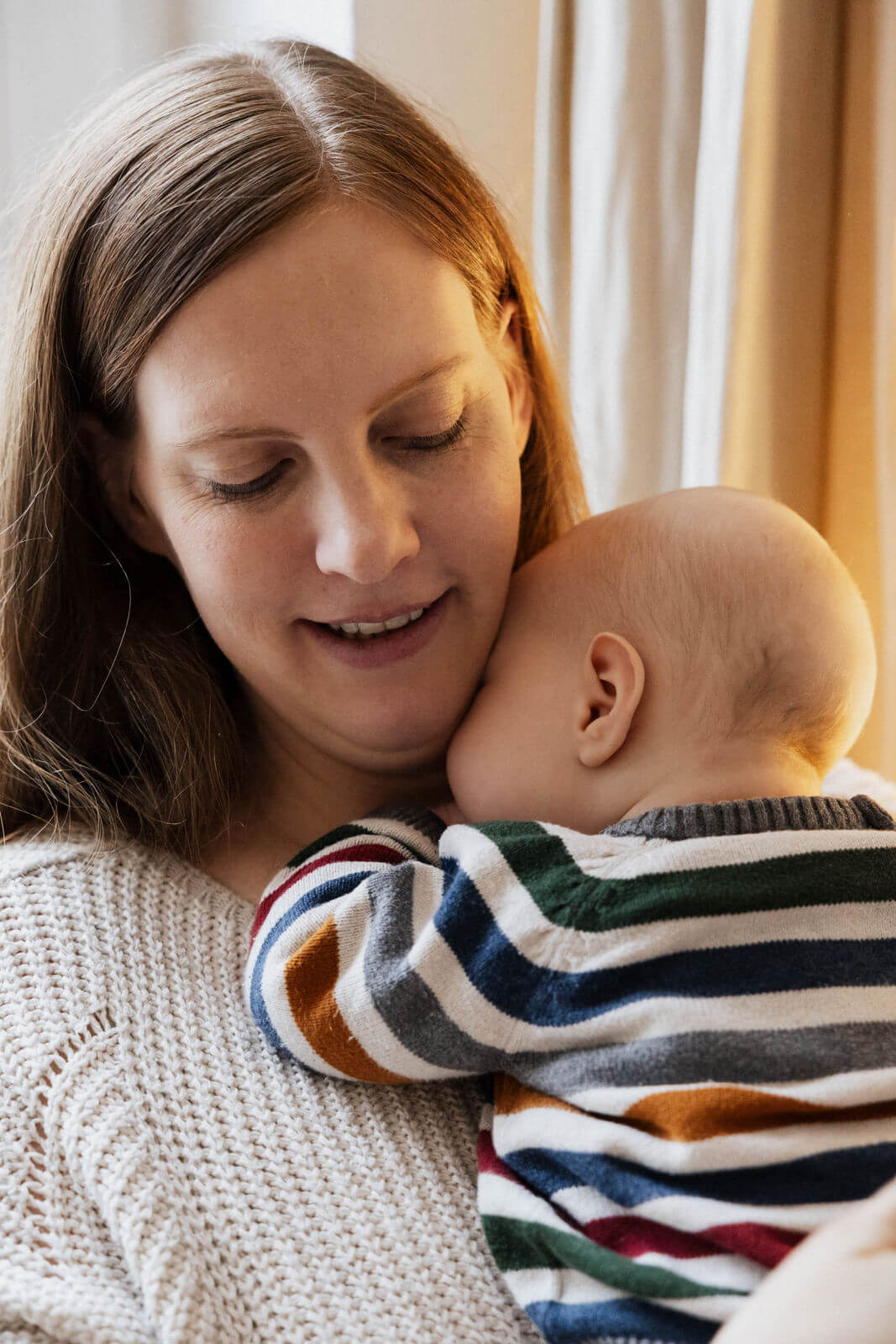 Mutter lächelt sanft, während ihr Baby im gestreiften Strampler zufrieden an ihrer Schulter liegt