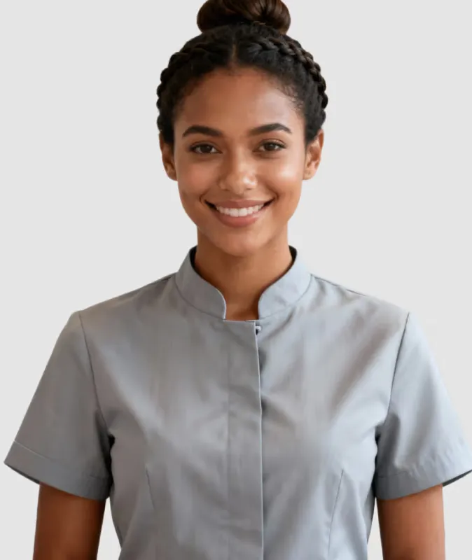 Smiling woman with braided hair bun wearing a gray short-sleeve uniform top against plain background.