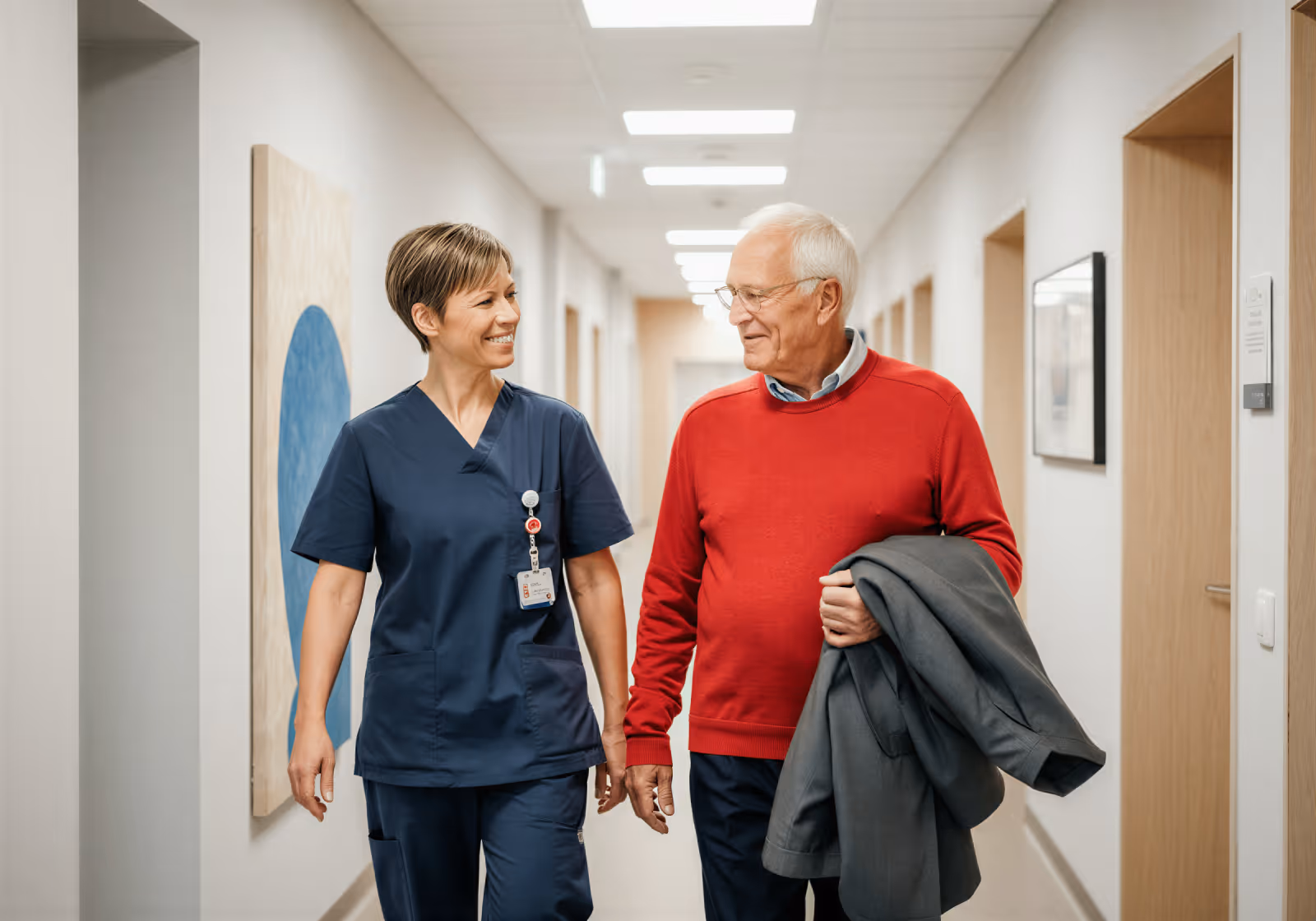 Smiling female nurse walking and talking with elderly man holding a coat in a hallway.
