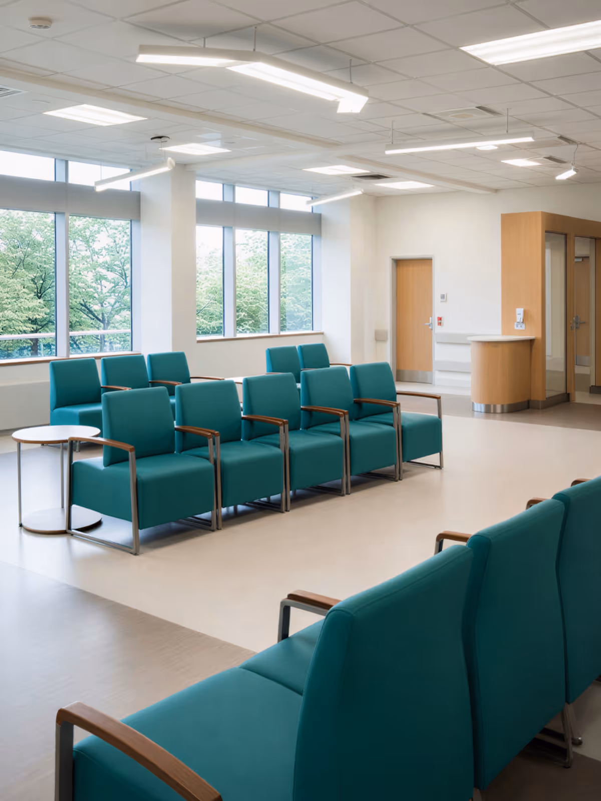 Modern waiting room with teal chairs and large windows showing green trees outside.