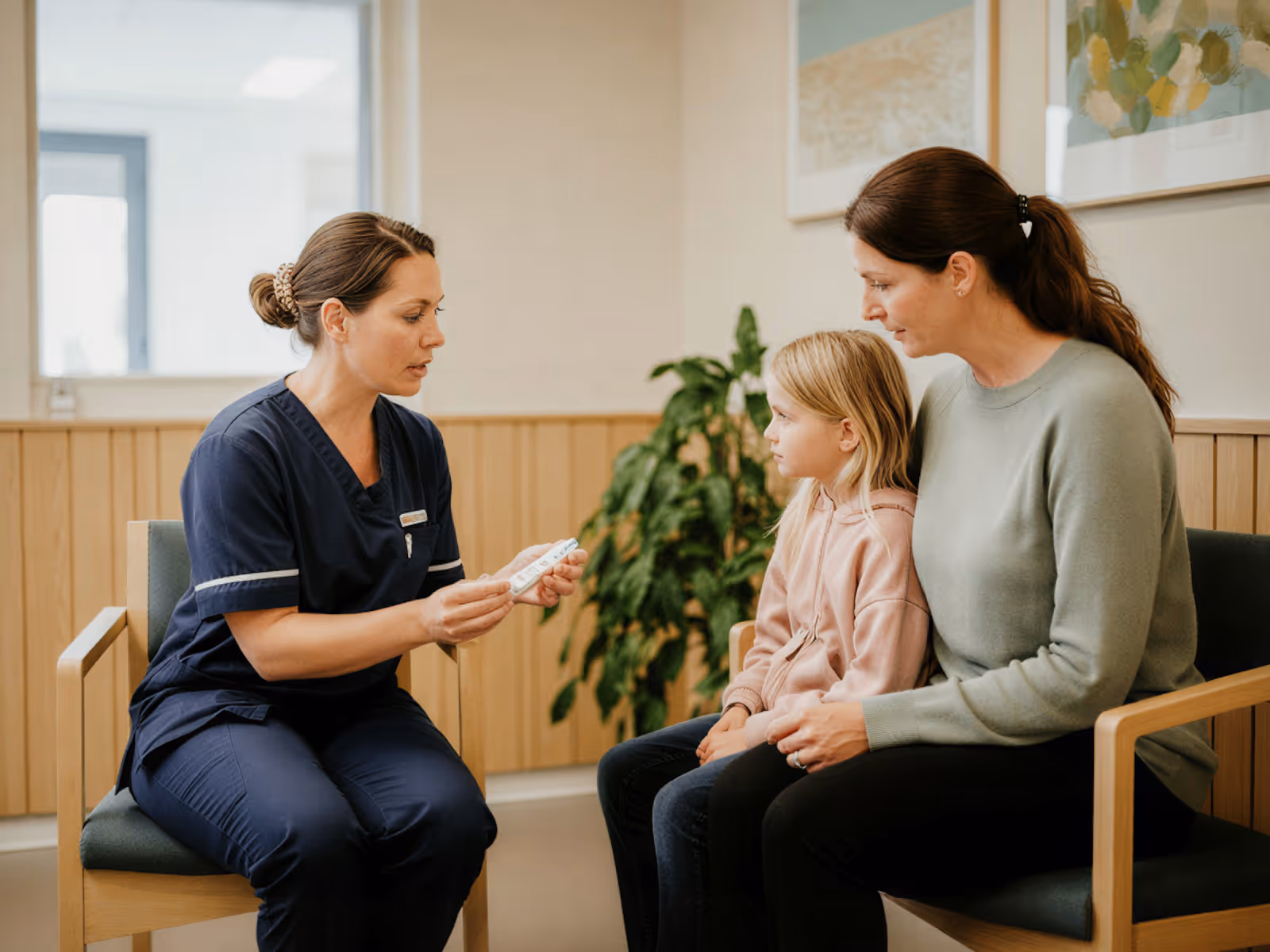 Nurse explains a medical test to a woman and child seated in a clinic waiting room.
